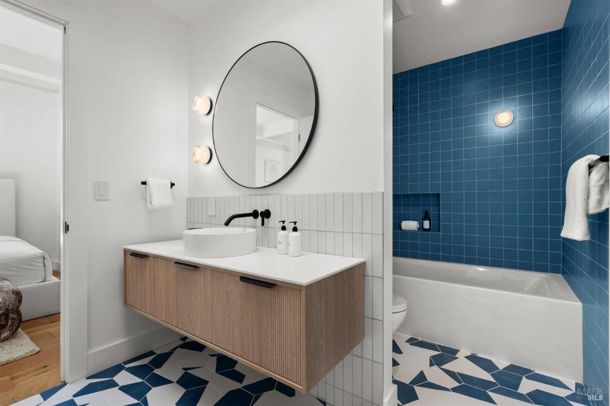 A bathroom with a round mirror, blue tiled shower, and a modern vanity.