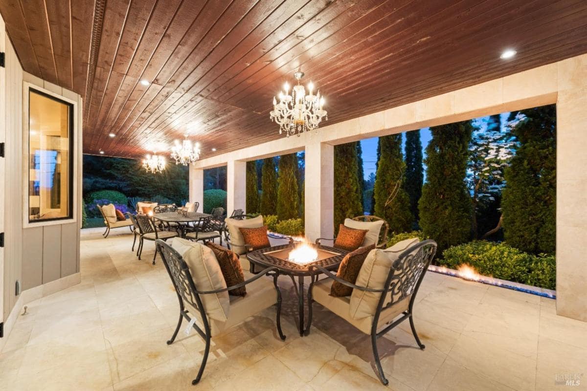 A covered patio with seating, a fire pit, and chandeliers. Large windows provide views of the outdoors.