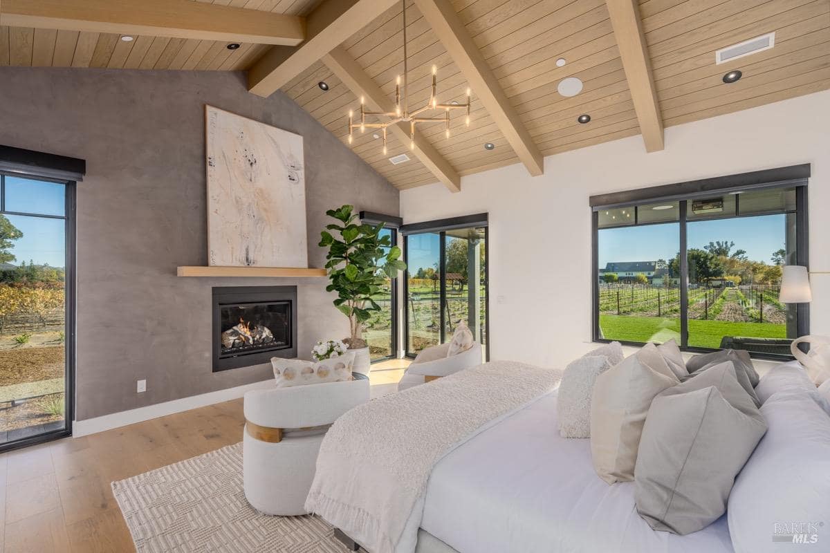 A spacious bedroom with a vaulted ceiling, fireplace, large bed, seating area, and windows overlooking the vineyard.