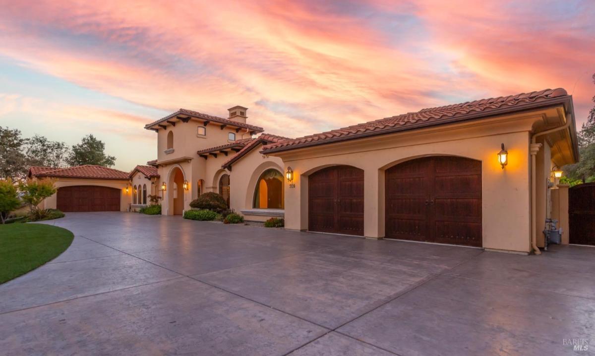 home with a tiled roof features a spacious driveway and three garage doors, set against a vibrant sunset sky.