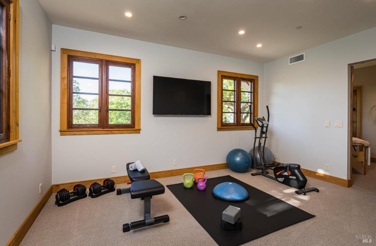 A home gym with a wall-mounted TV, windows, and fitness equipment on the floor.