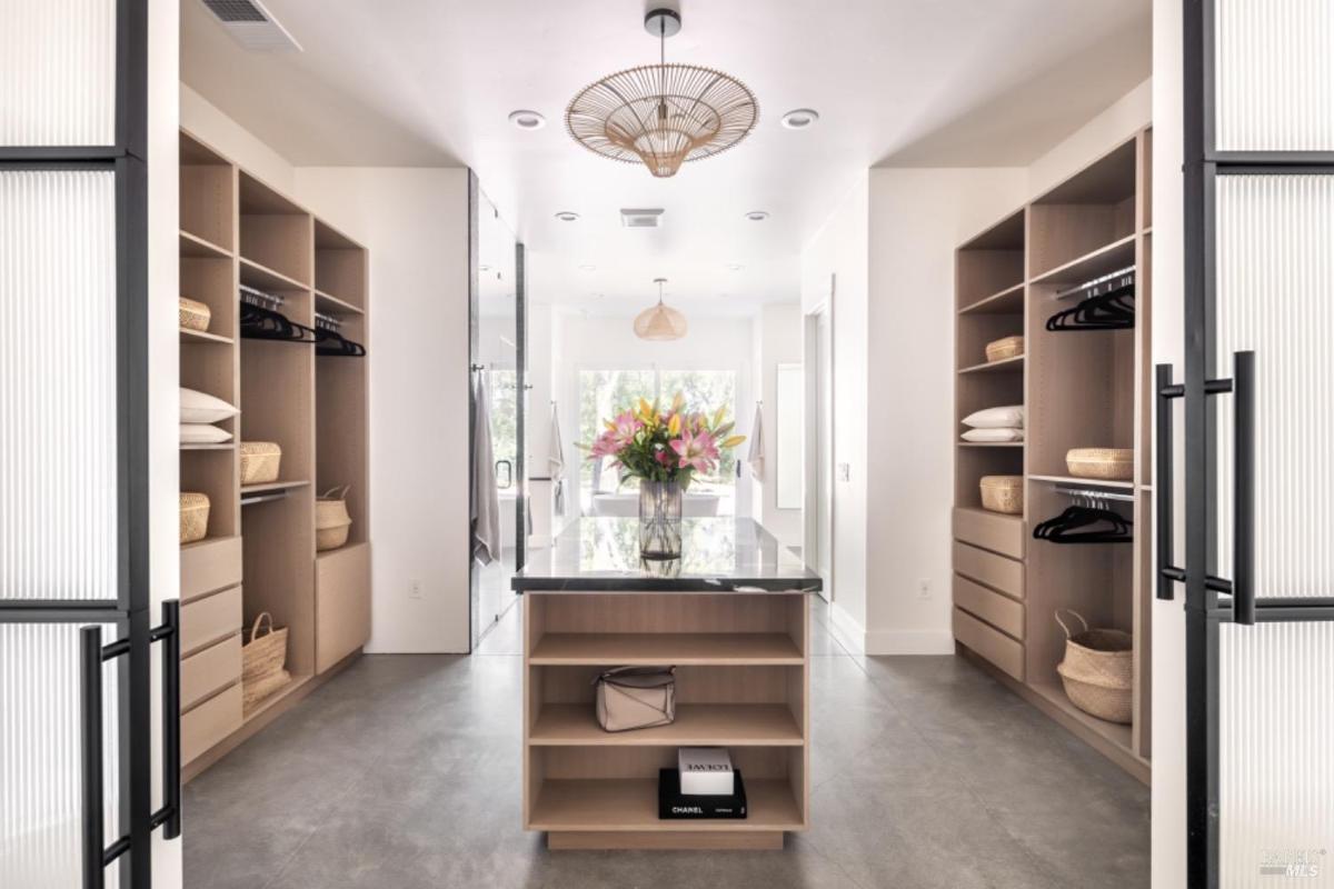 A walk-in closet with open shelving, a central island with a glass top, and modern lighting fixtures.