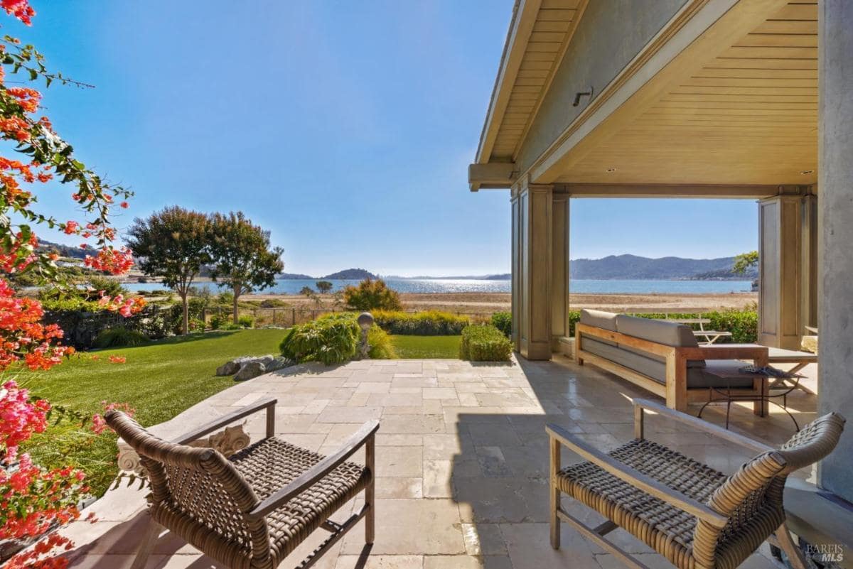 An outdoor living area featuring stone flooring and comfortable seating, framed by vibrant flowers and a stunning view of a lake and hills in the background.