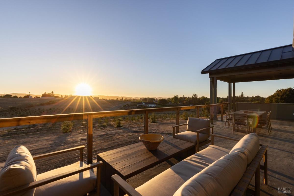 A wooden deck with a dining table and chairs faces a sunset over a grassy landscape.