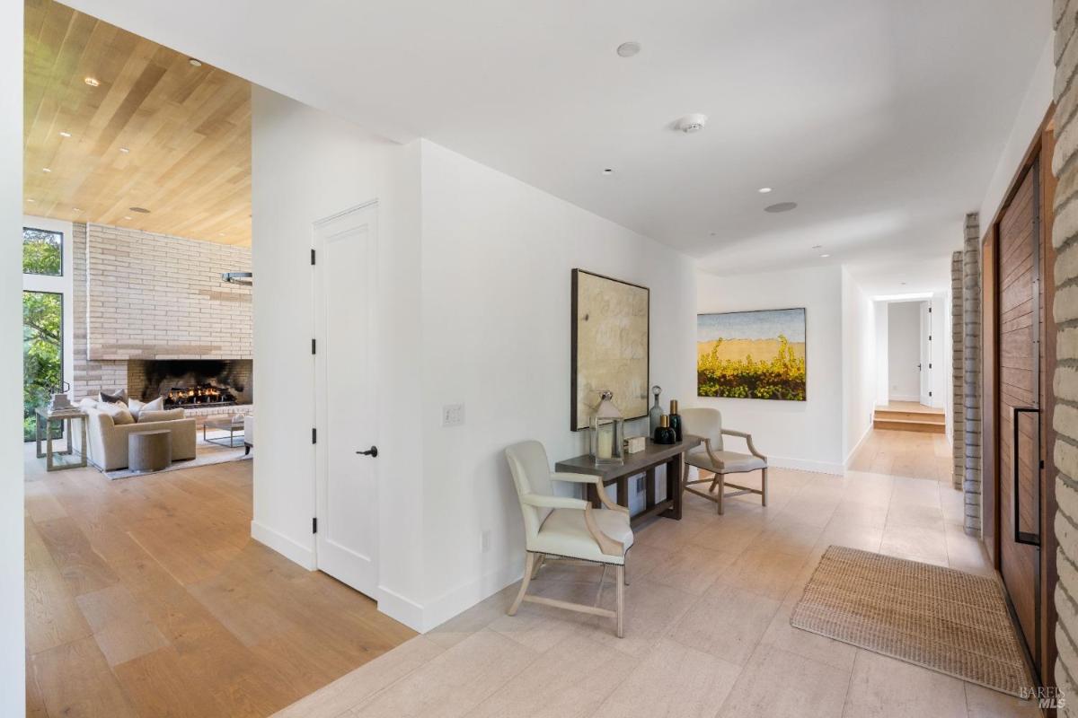 A foyer view with light-colored flooring, white walls, artwork, a small console table, and chairs. A doorway leads to a living area with a fireplace.