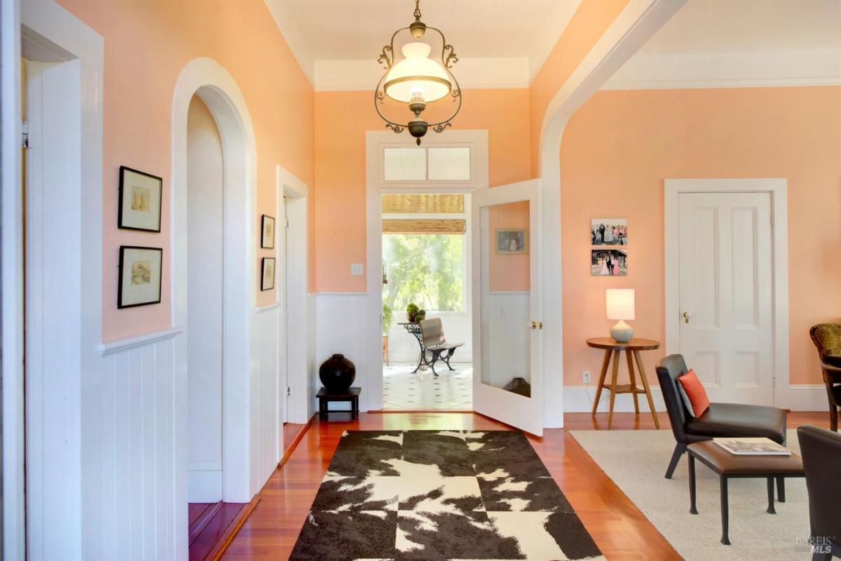 A foyer with peach-colored walls, white paneling, and a light fixture, leading to a glass door opening into a tiled sunroom.