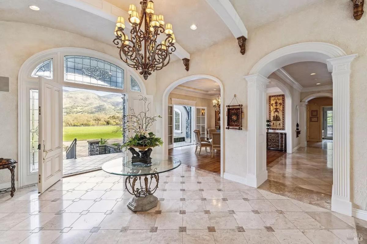 An entry with a tiled floor, a central glass table, and a chandelier. Archways lead to adjacent rooms.