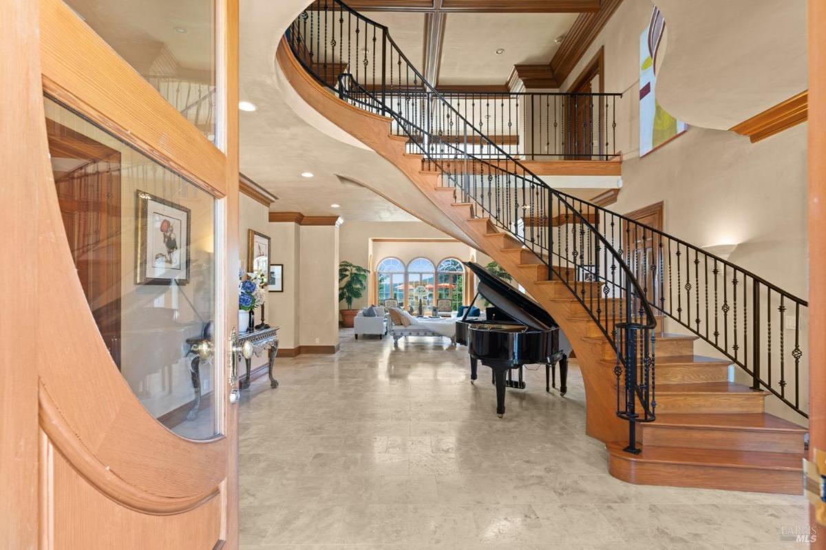 Foyer with sweeping staircase, polished floors, and a grand piano, leading to an open living space.