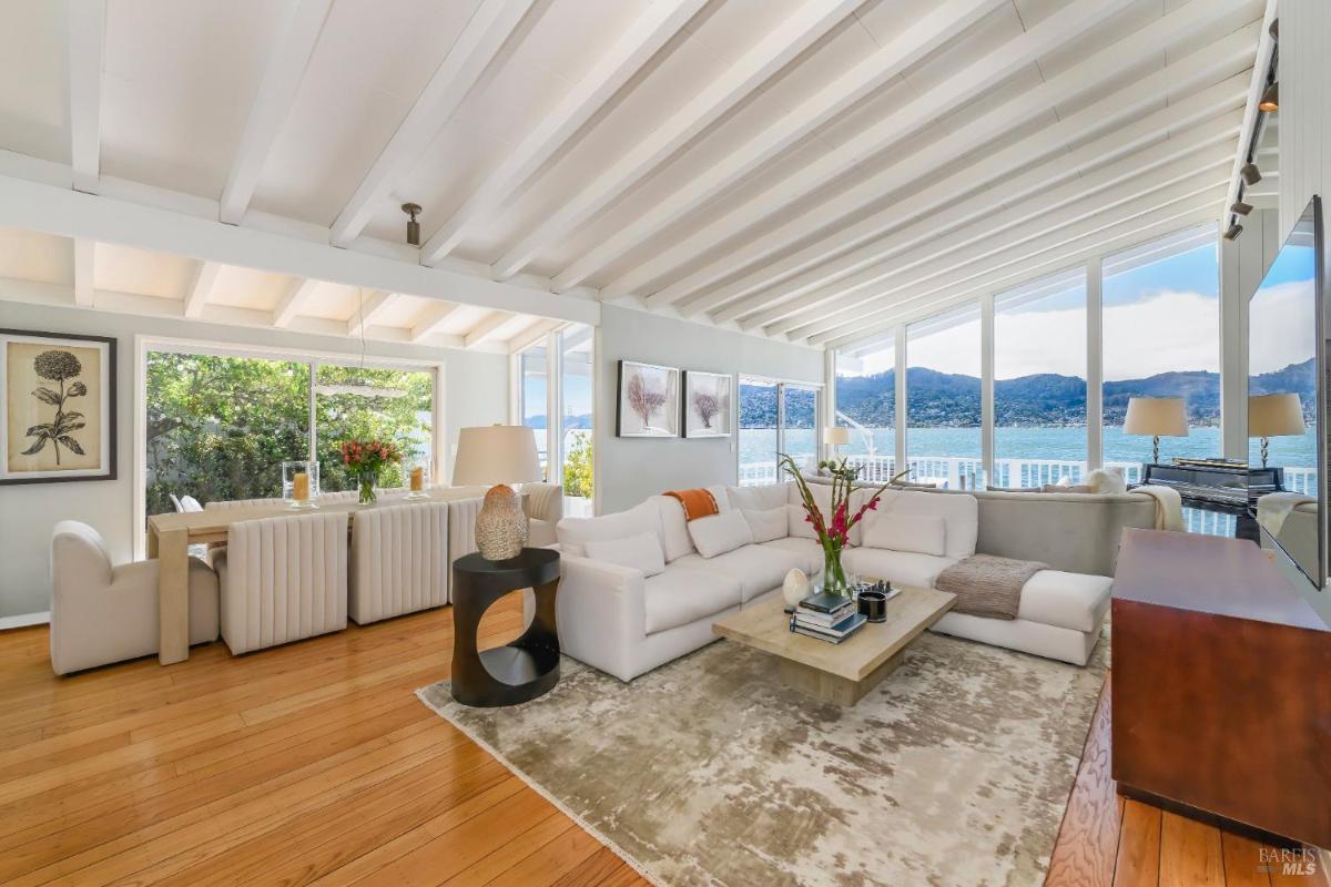 Open living and dining area with white furniture, wooden flooring, and views of water and greenery.