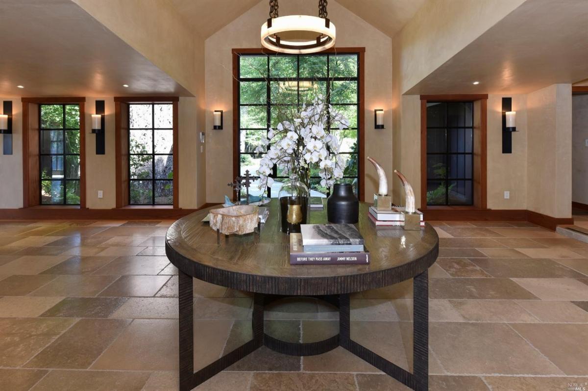 A foyer with round table with decor and books in the center of a room with large windows and stone flooring.