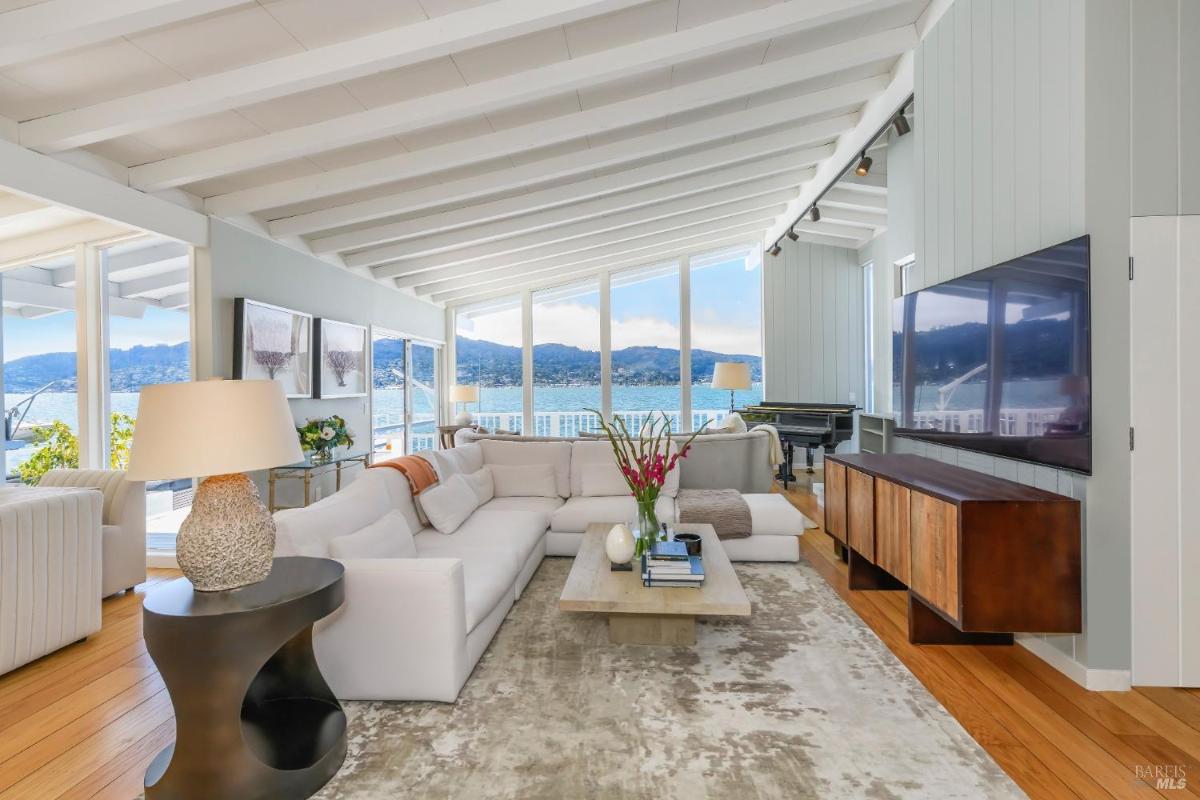 Living room with large sectional couch, wooden floors, and a view of the water through floor-to-ceiling windows.