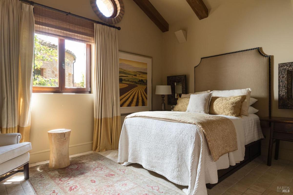 A bedroom with a neatly made bed, decorative pillows, a wooden stool, and a large window letting in natural light. 