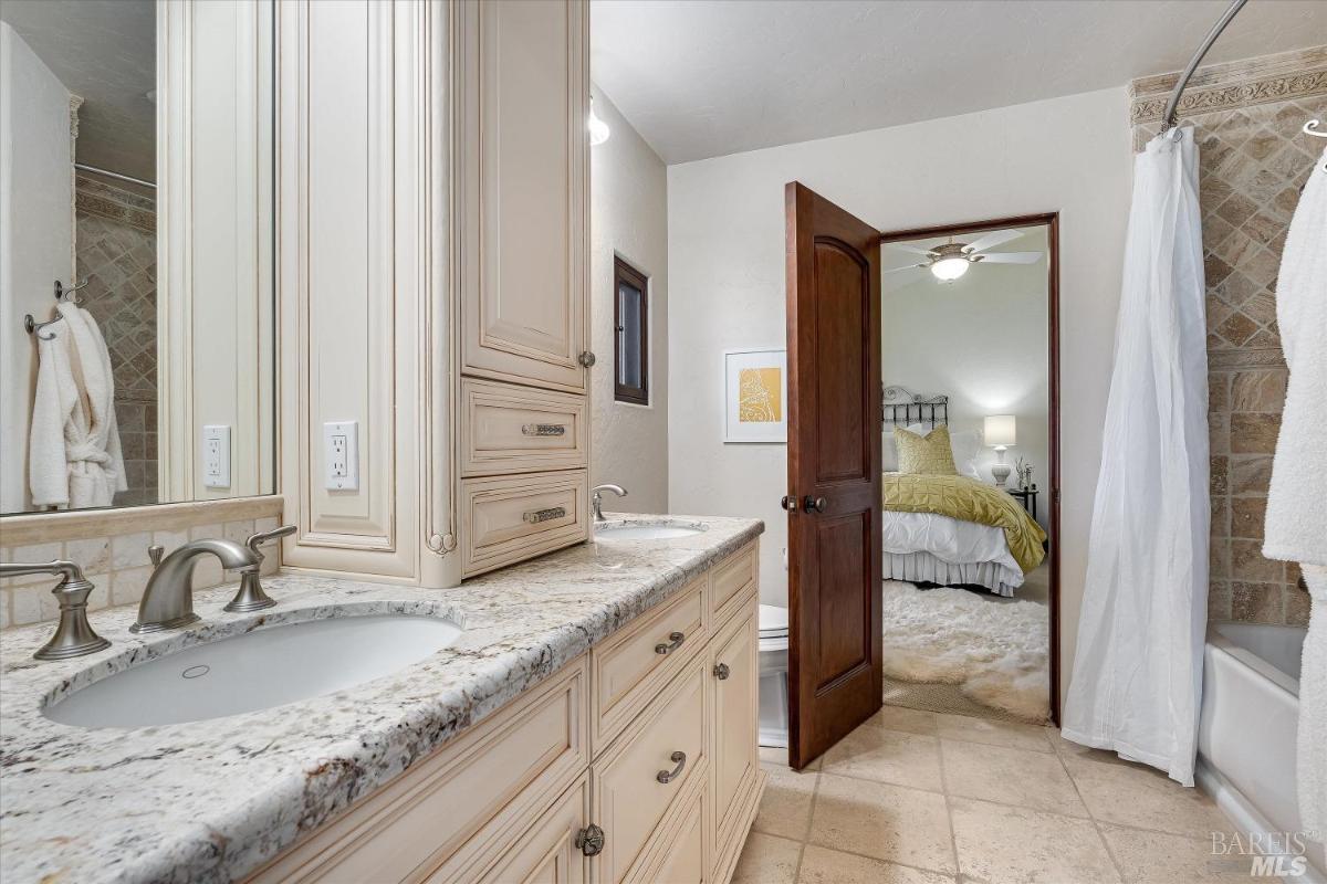 A bathroom with white cabinetry, granite countertops, and access to a bedroom.