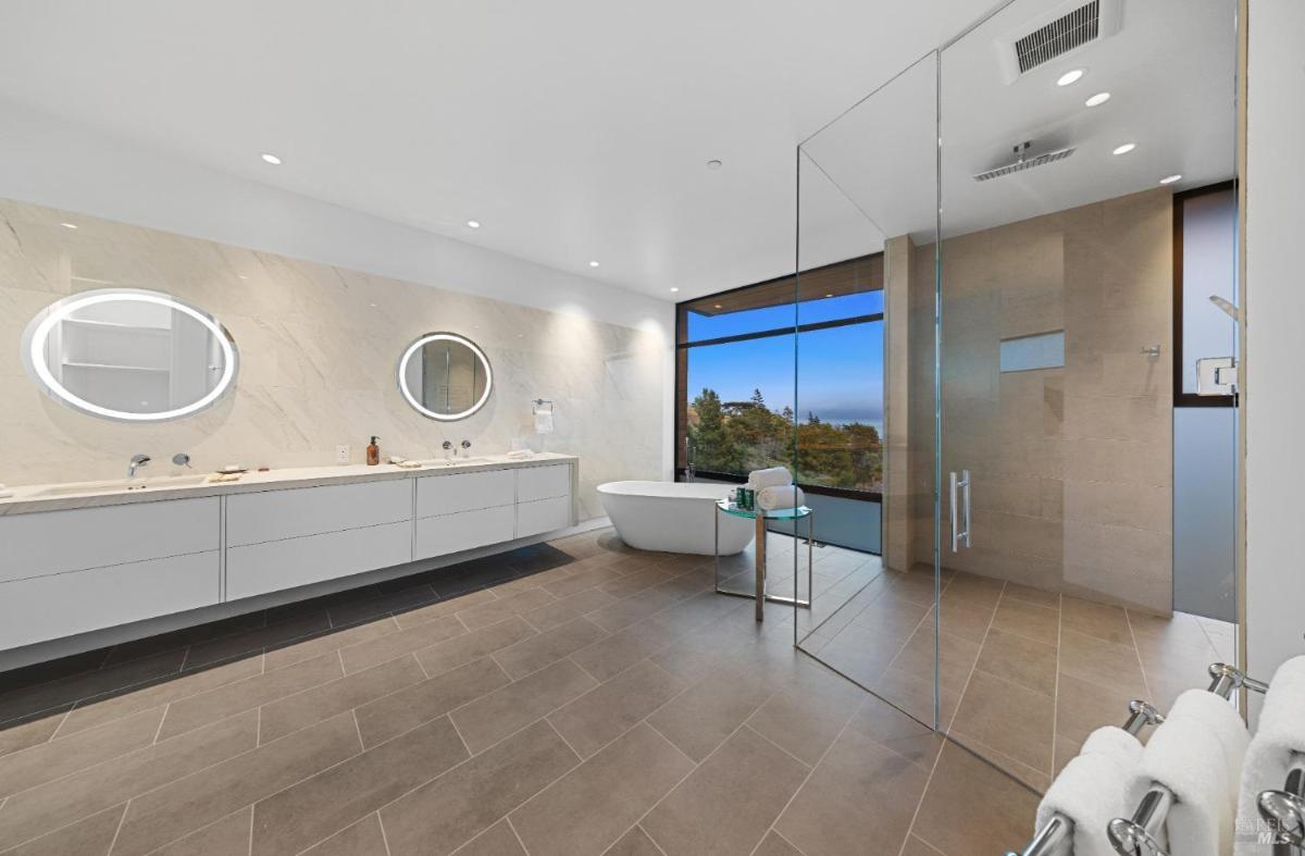 A spacious bathroom with a double vanity, circular mirrors, a freestanding tub, and a glass-enclosed shower overlooking a hillside.