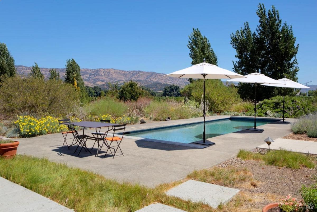 A concrete patio with a rectangular pool, a dining table, and white umbrellas, surrounded by shrubs and distant hills.