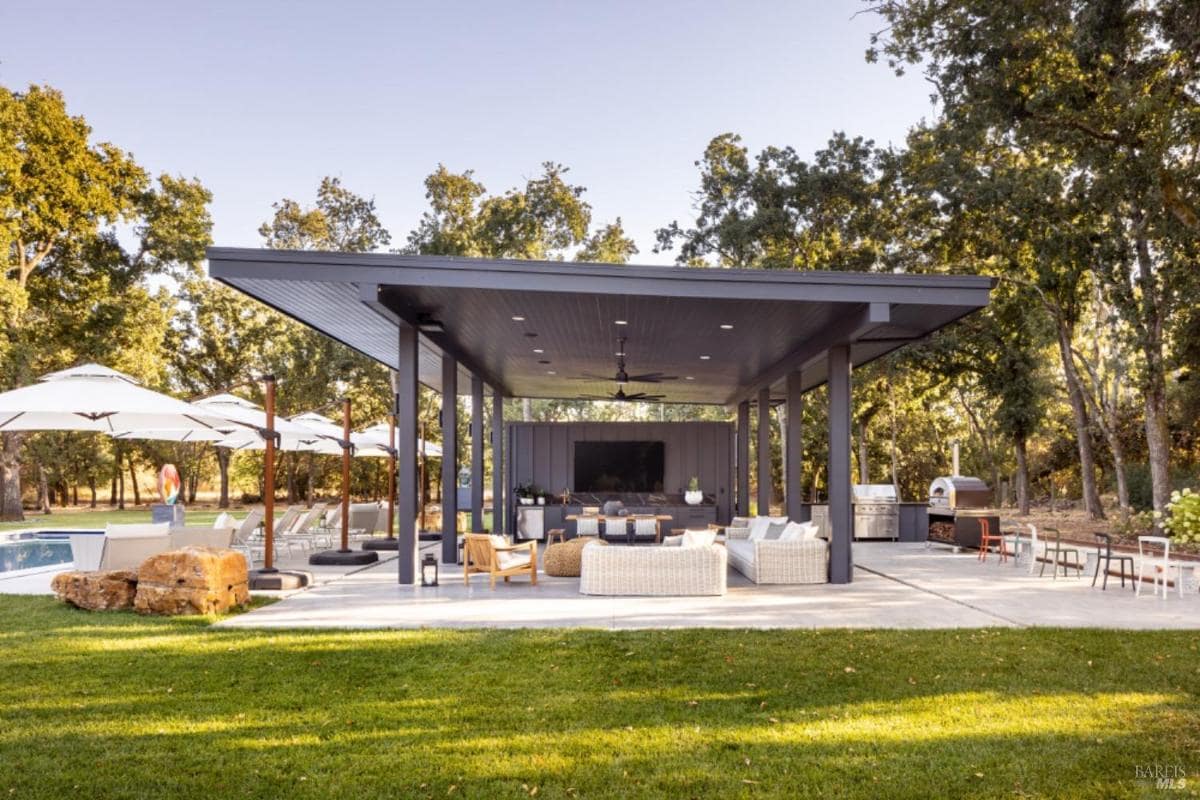 An alfresco kitchen with a covered seating area, large TV, grilling station, and poolside lounge chairs.