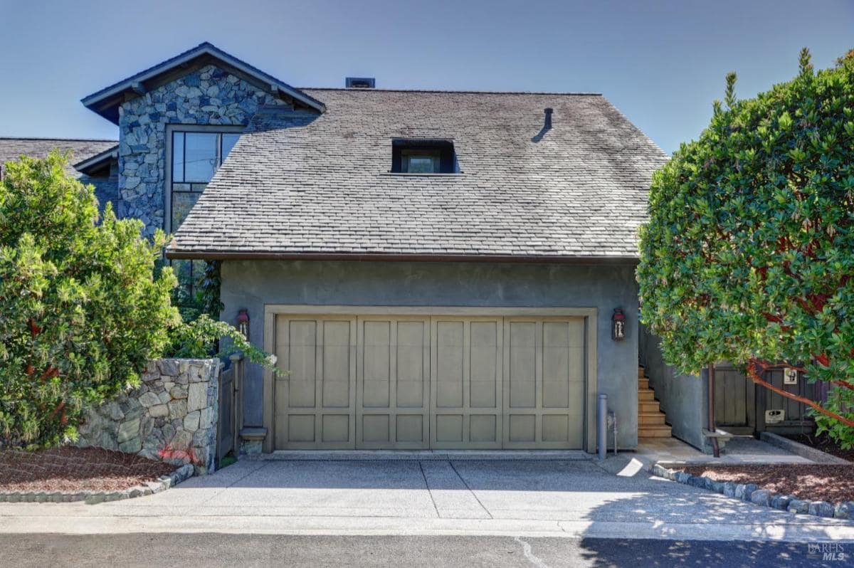 Garage with stone exterior and a sloped roof, featuring a spacious garage door and lush greenery surrounding the entrance. 