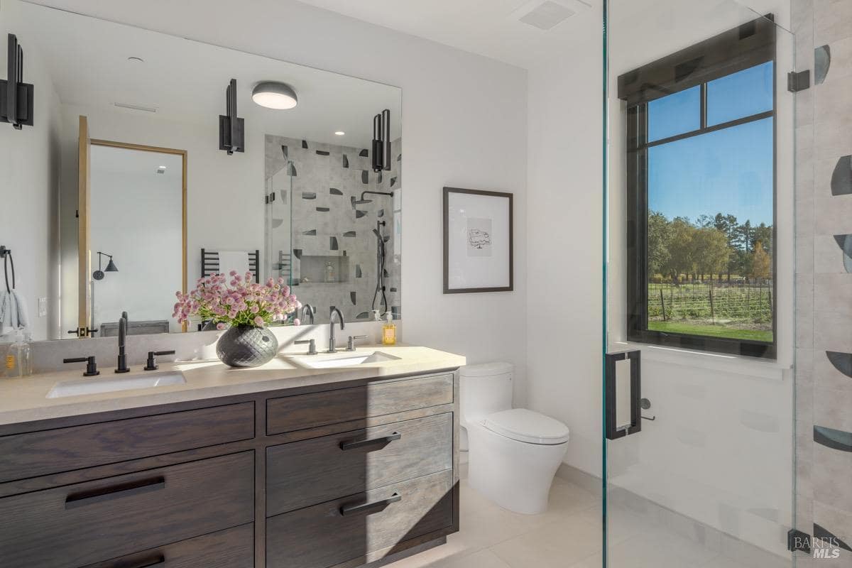 A bathroom with a double vanity, walk-in shower, toilet, and a large window overlooking the vineyard.