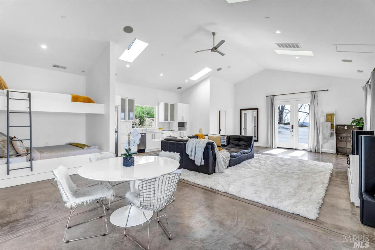 A spacious room featuring a lofted bed, a dining table, and a cozy seating area, with large windows and a modern aesthetic.