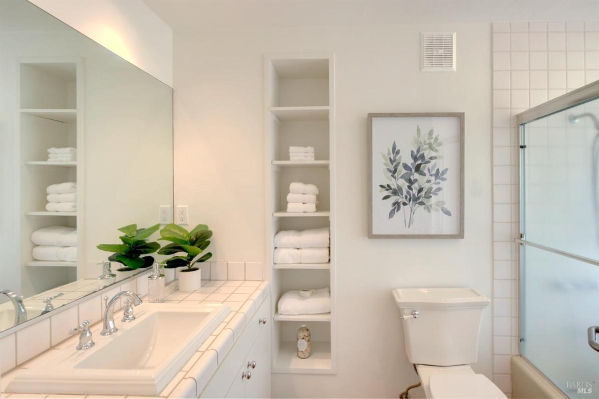 A bathroom with a tiled sink and built-in shelving, featuring a framed plant artwork.