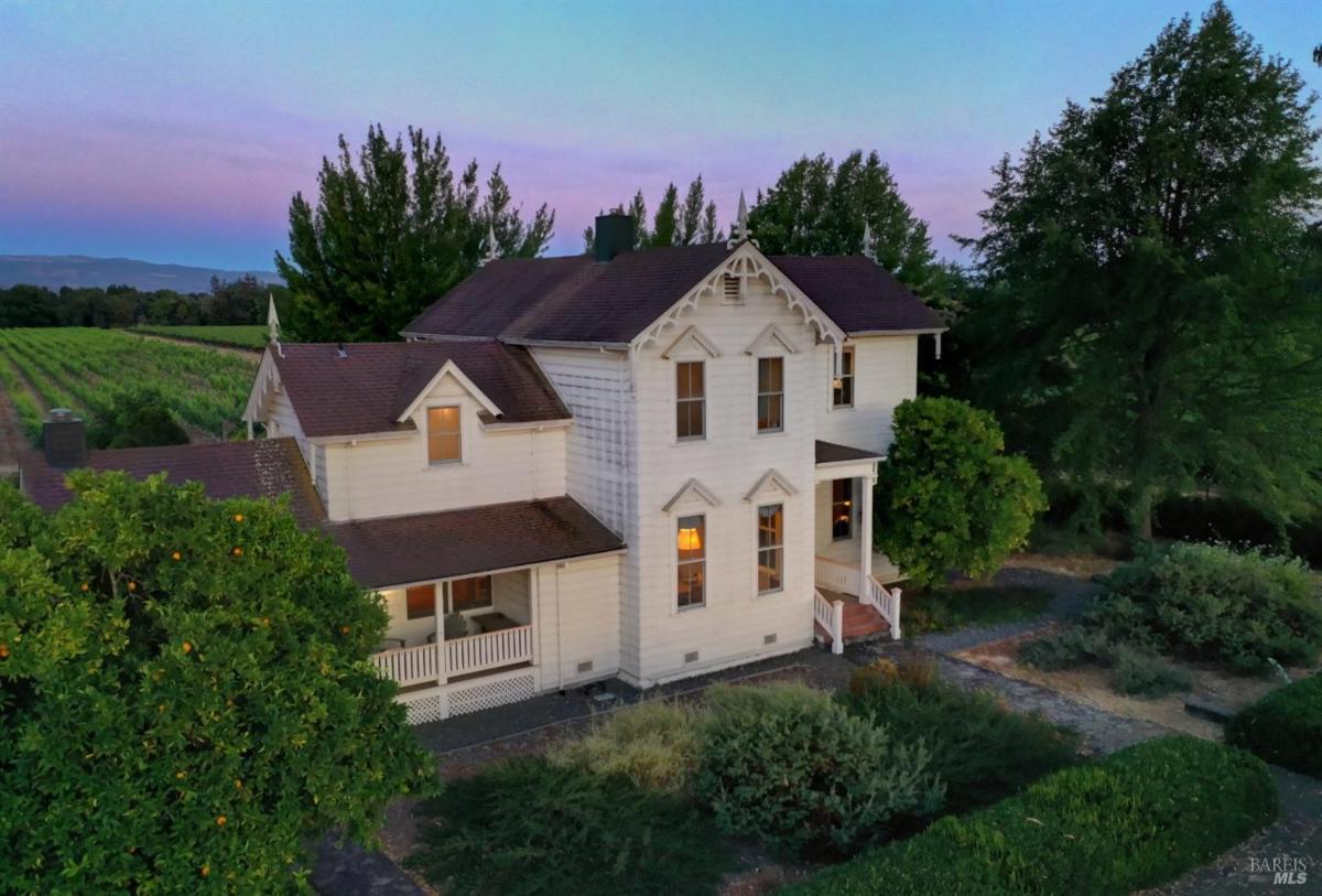 A large white house with a porch, surrounded by trees and vineyards, with a sunset sky in the background.