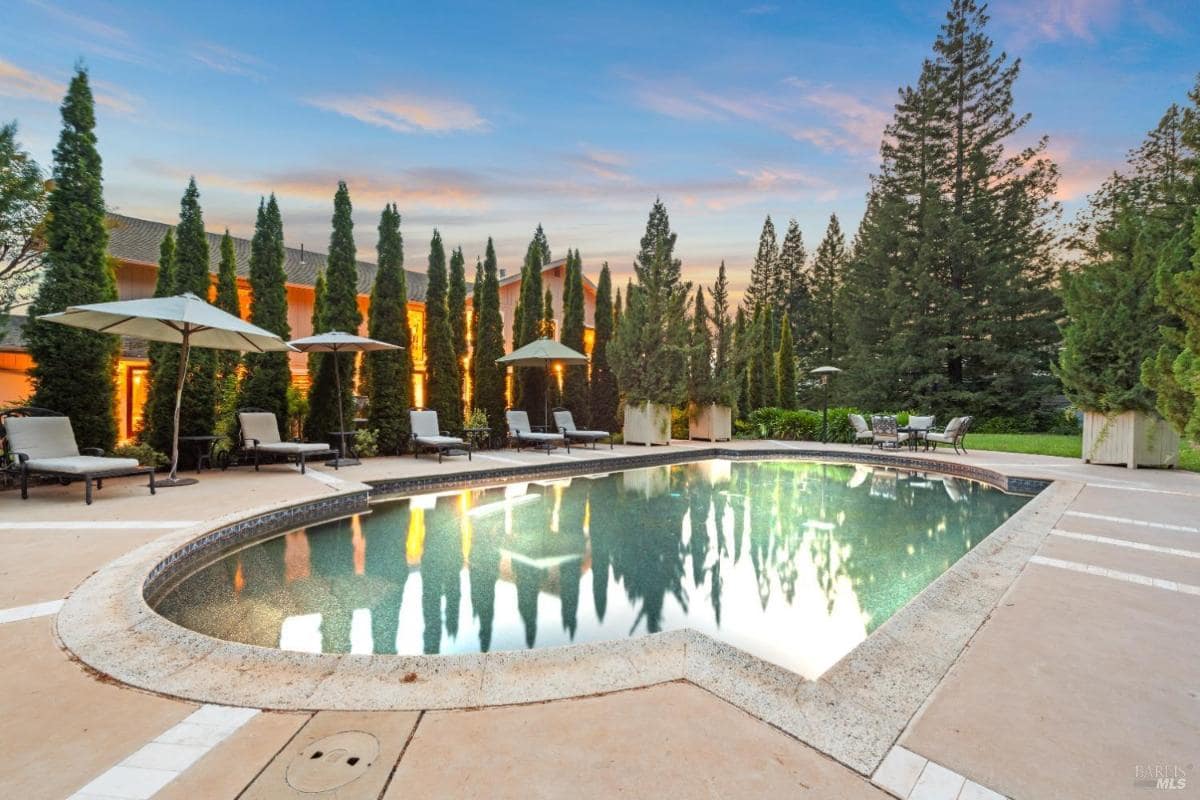 A swimming pool surrounded by lounge chairs and tall trees, with umbrellas nearby.