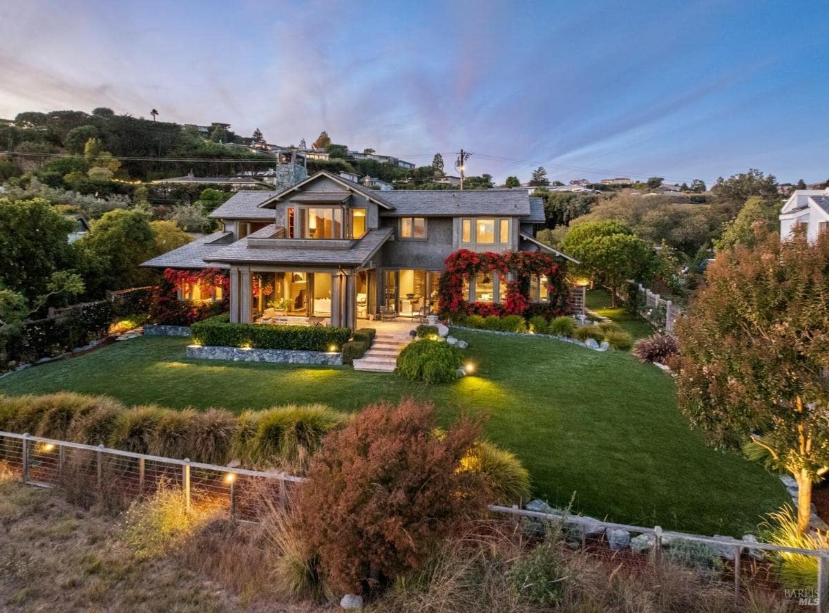 A house surrounded by lush gardens and vibrant flowers, illuminated by soft outdoor lighting at dusk. 