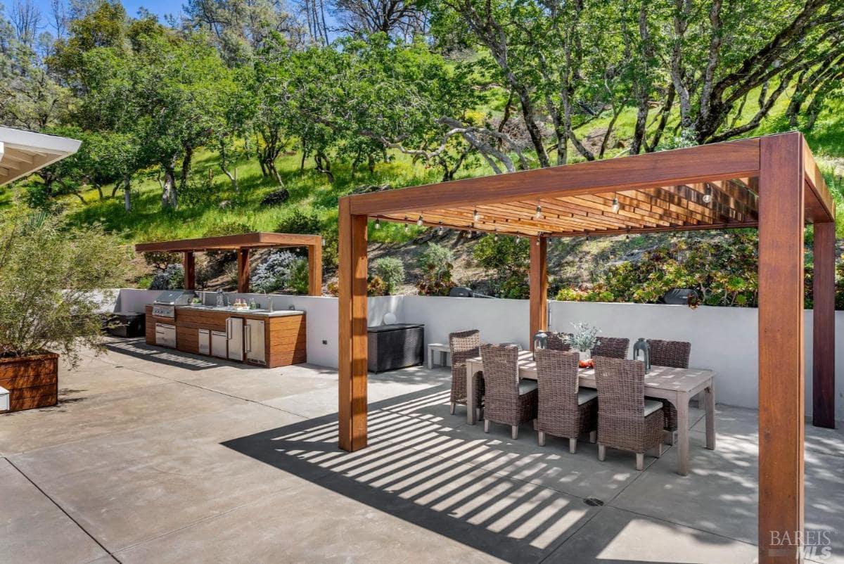 An outdoor space with wooden pergolas, dining furniture, and a barbecue area, surrounded by greenery and a hillside backdrop.