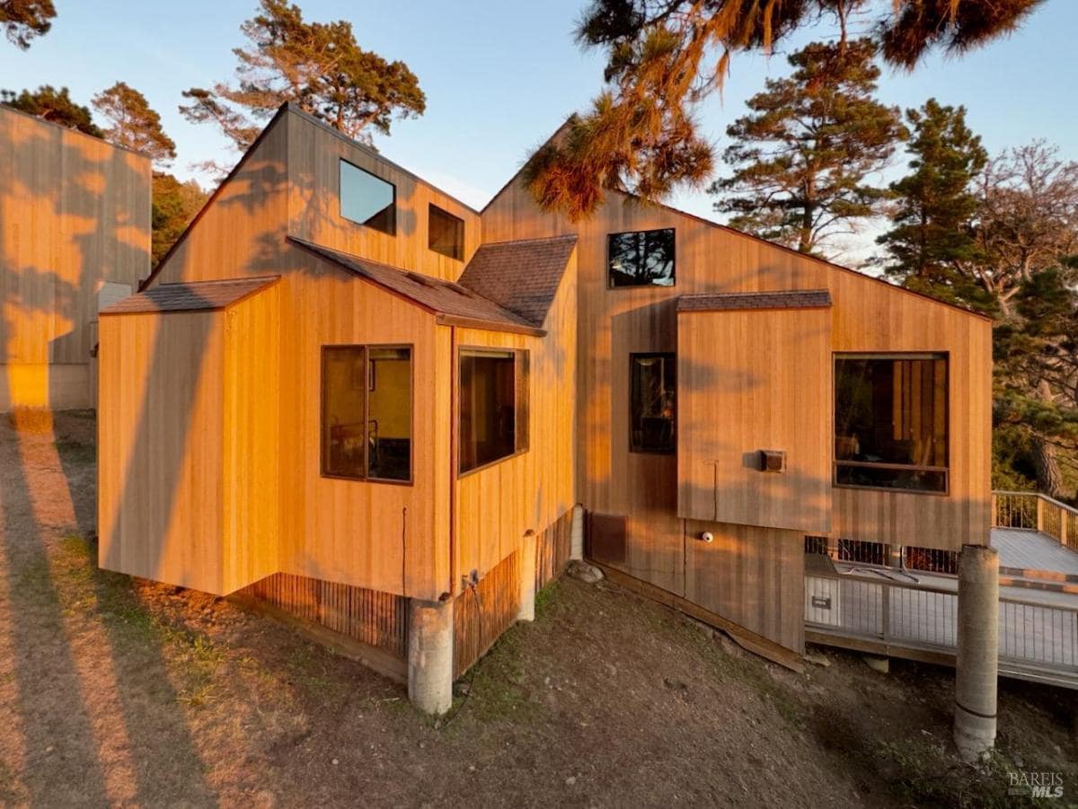 A modern wood-clad home with large windows reflecting sunlight, set on a hillside. 