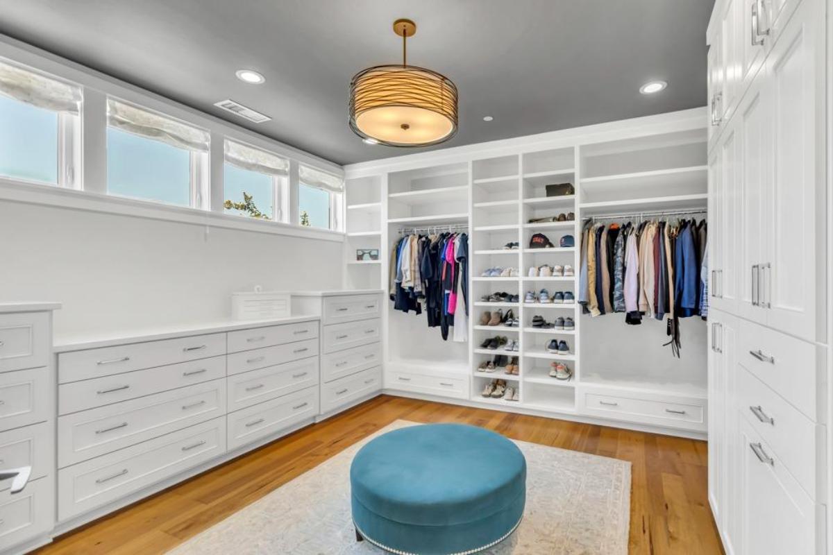 A walk-in closet features white cabinetry, shelves for shoes, and a blue ottoman.