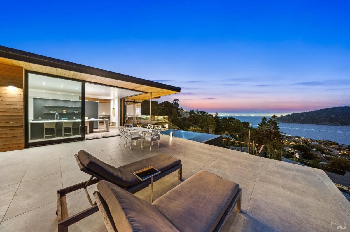 A patio with lounge chairs, a dining table, an infinity pool, and a bay view.