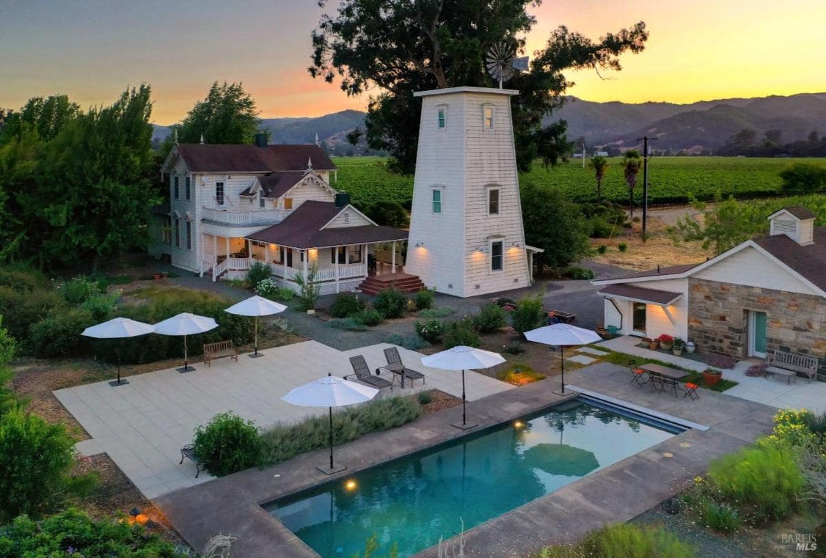 A white house, a tower, and a swimming pool with umbrellas and patio furniture, surrounded by greenery and vineyards, under a sunset sky.