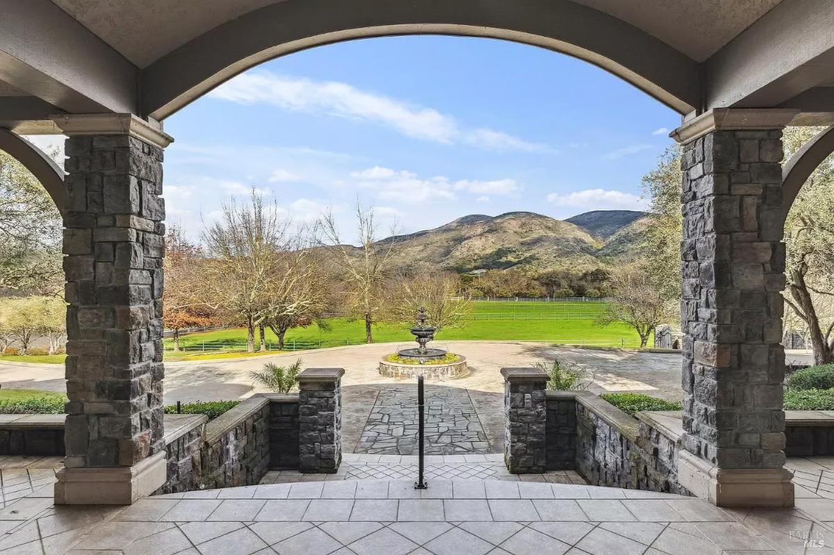 Outdoor view from an arched patio overlooking a circular driveway, fountain, and a green field with hills in the background.