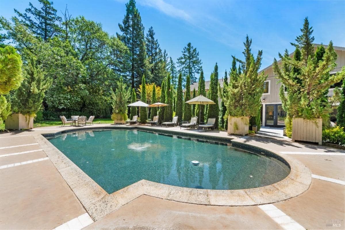 A swimming pool surrounded by lounge chairs and tall trees, with umbrellas nearby.