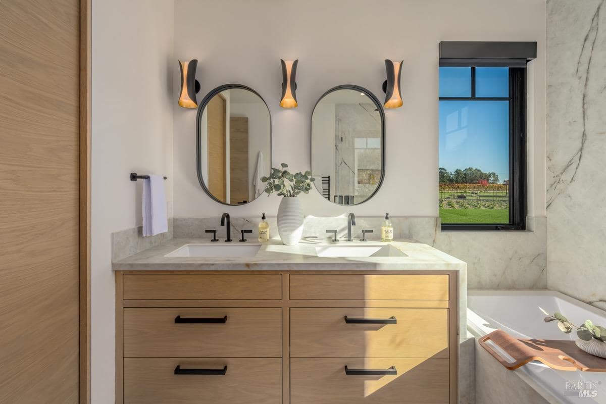 A bathroom with a double vanity, two mirrors, modern lighting, and a built-in bathtub.