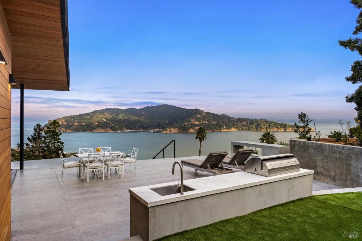 A patio area with a dining table, lounge chairs, a built-in grill, and a bay view.