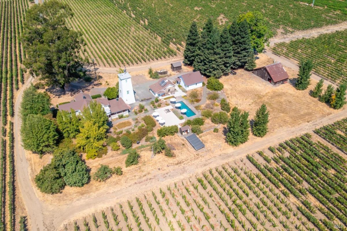 An aerial view of a mansion featuring a white house, tower, pool, outbuildings, and surrounding vineyards.