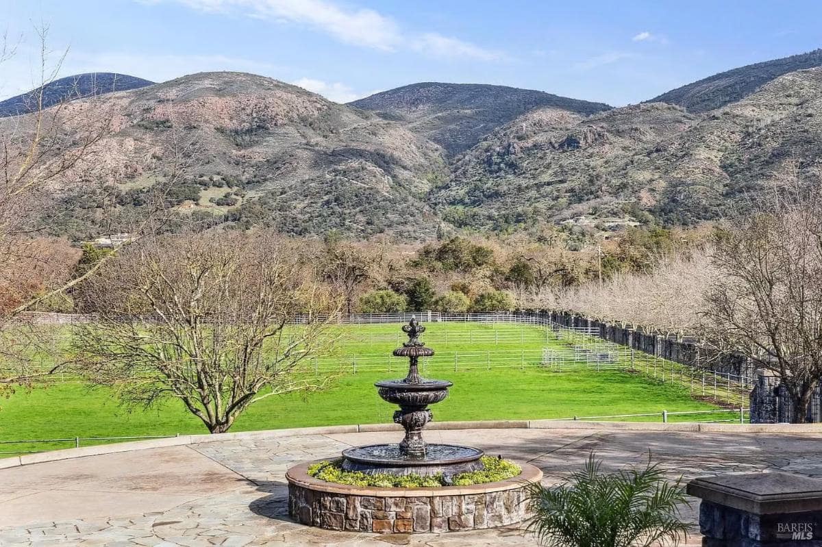 Outdoor shot of a fountain in a circular driveway surrounded by trees, with hills in the background.