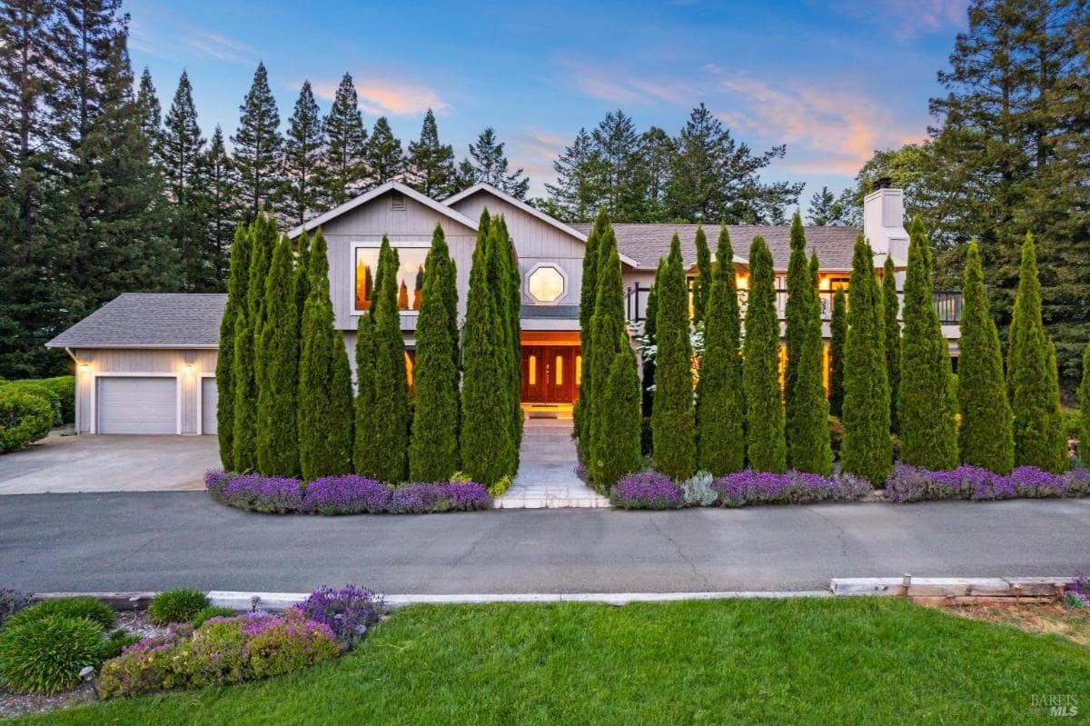 A large house with a circular window, surrounded by tall trees and colorful flowers along the driveway.