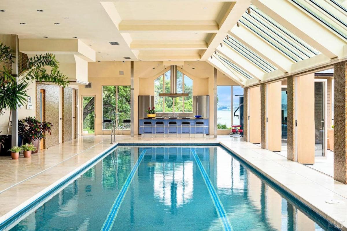 Indoor pool area with a modern kitchen, vaulted ceilings, and expansive skylights. Sliding glass doors provide access to outdoor patios and views.