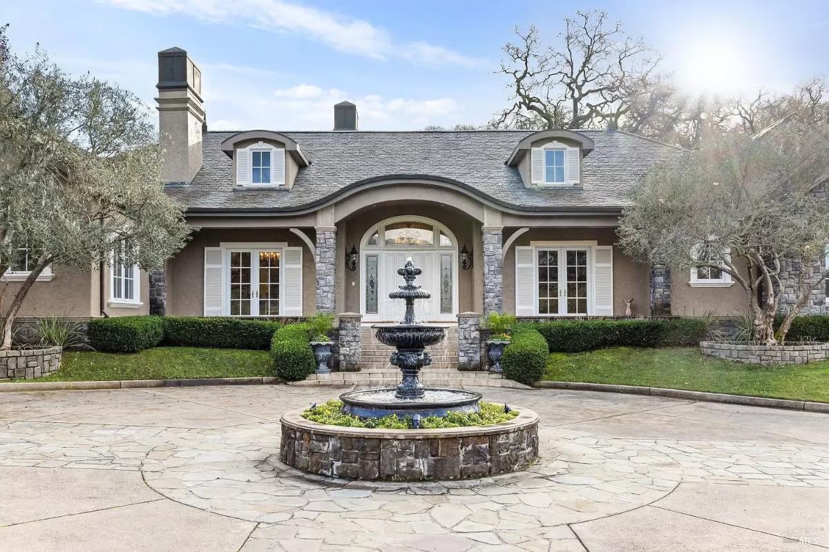 Front view of a house with a central entrance, stone and stucco exterior, and a circular driveway with a fountain.