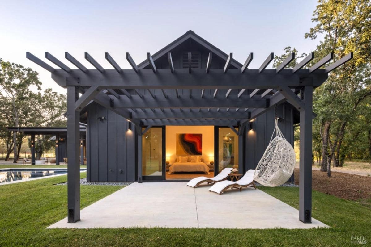 A pergola with a hanging chair, two lounge chairs, and large sliding doors leading to a bedroom.