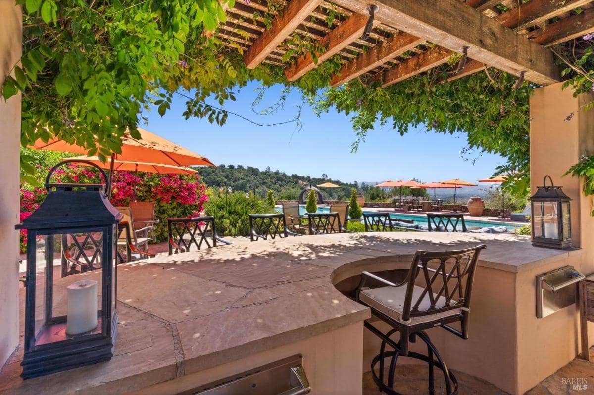 Bar-style outdoor kitchen under a pergola with vibrant plants and pool views.