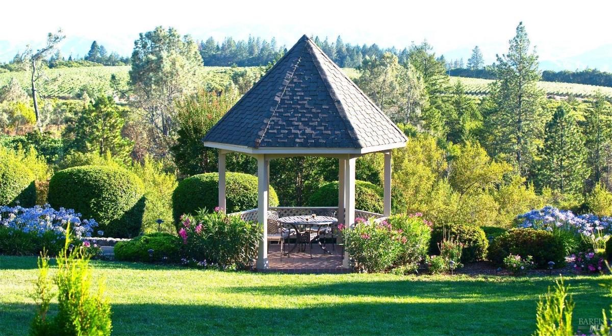 A gazebo in a lush garden with neatly trimmed bushes and colorful flowers, overlooking a scenic landscape of trees and hills.