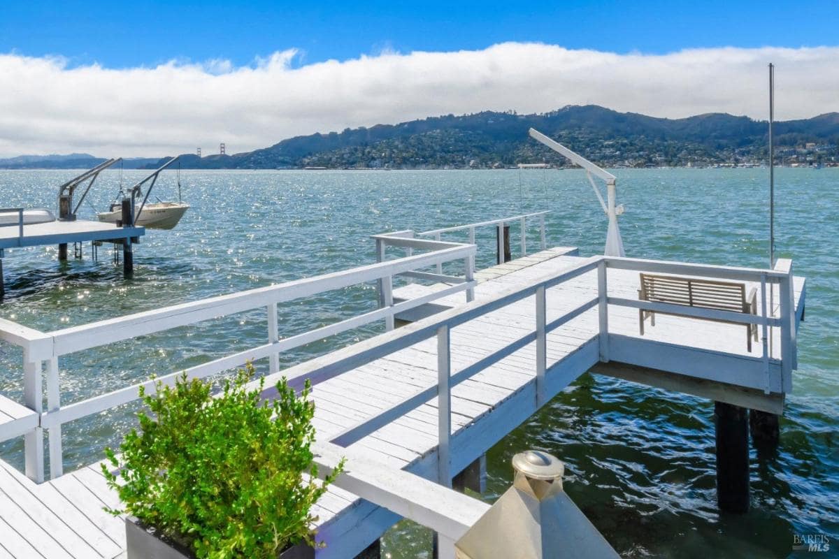 A white dock extending over the water, equipped with a bench and boat lifts, with hills in the background.