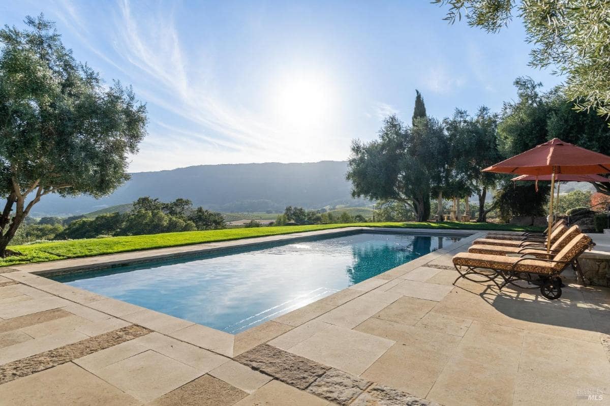 A rectangular outdoor pool with lounge chairs and umbrellas on the deck, surrounded by a landscaped lawn and trees.