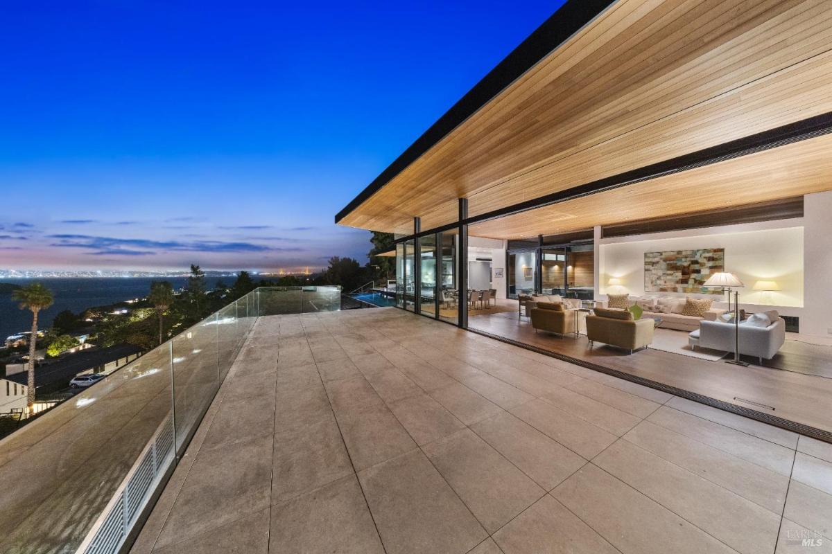 A deck with glass railing adjacent to a living area, overlooking a bay and city lights at night.