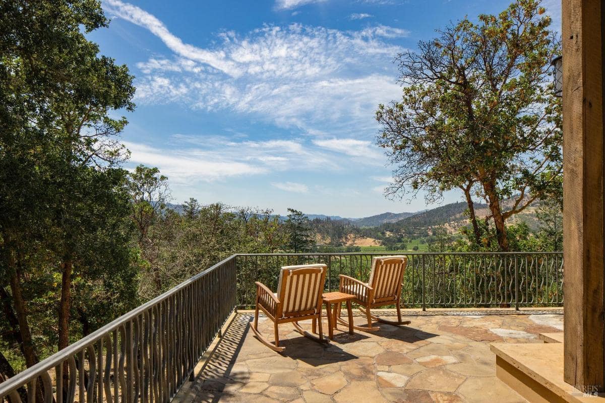 A stone patio with two wooden chairs overlooking trees and hills.