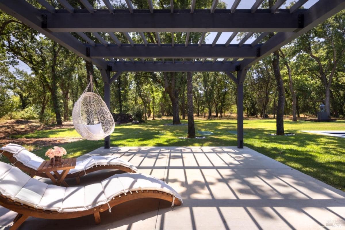 A pergola with a hanging chair, two cushioned lounge chairs, and a small table, surrounded by a shaded grassy area.