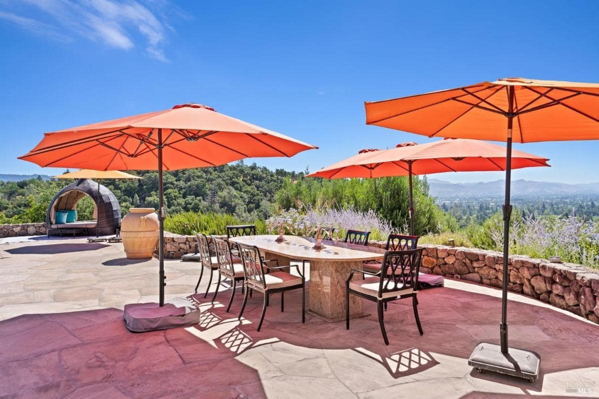 A spacious stone patio features a large dining table surrounded by eight chairs and shaded by vibrant orange umbrellas. 